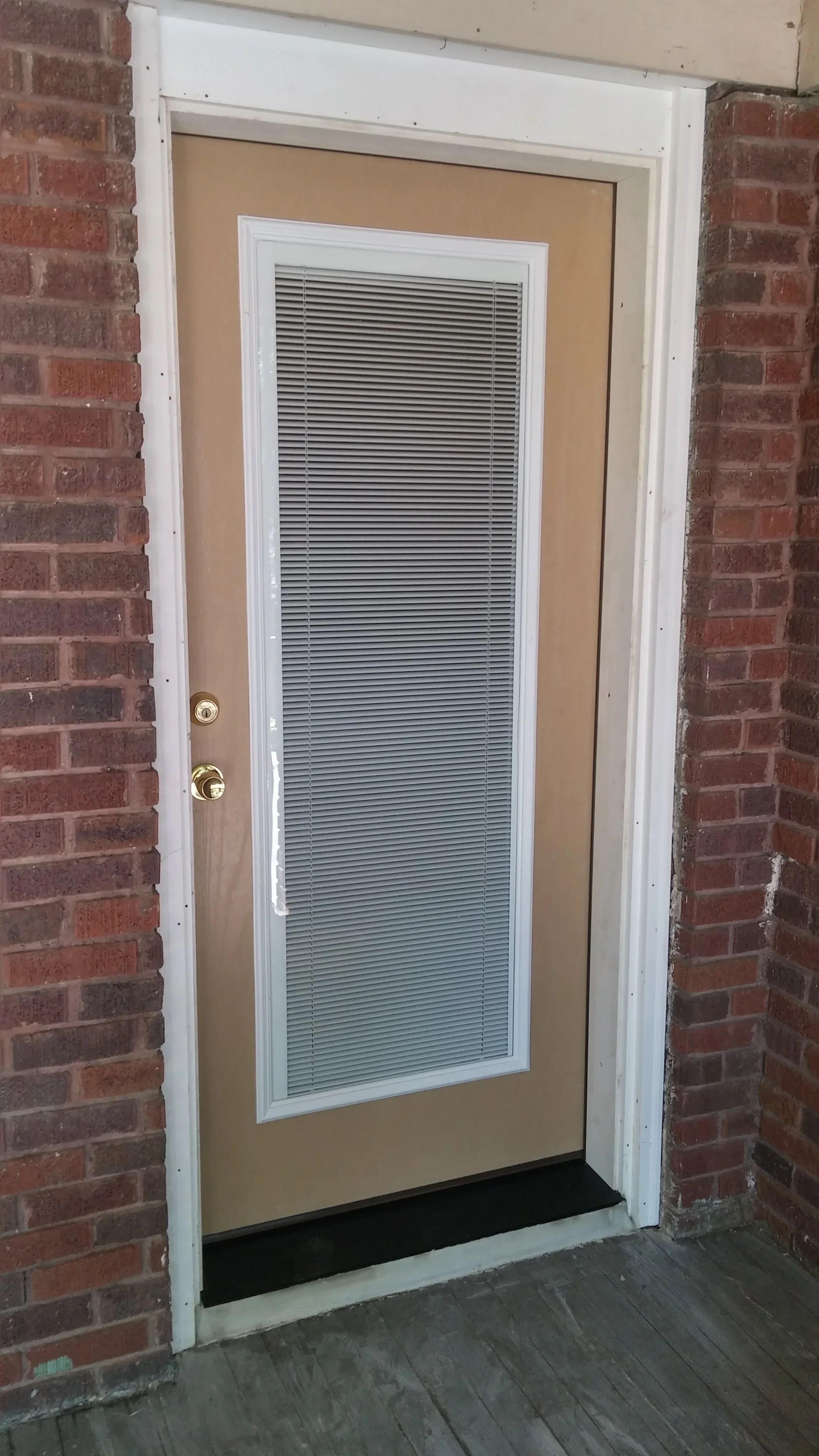 Full-glass door with integrated blinds in white frame and tan panel between brick walls
