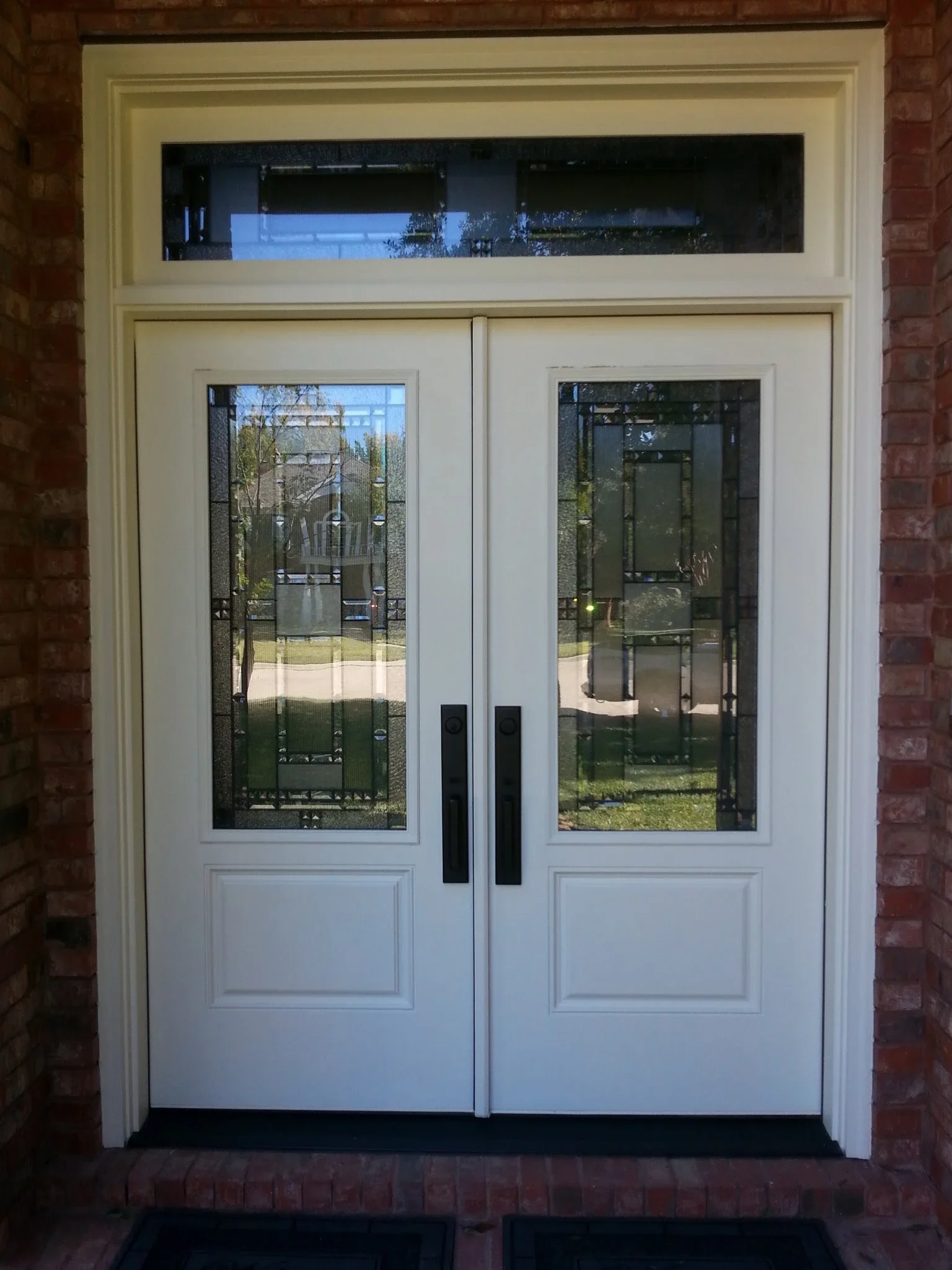 White aluminum double doors with glass panels and black handles, transom window above, brick exterior