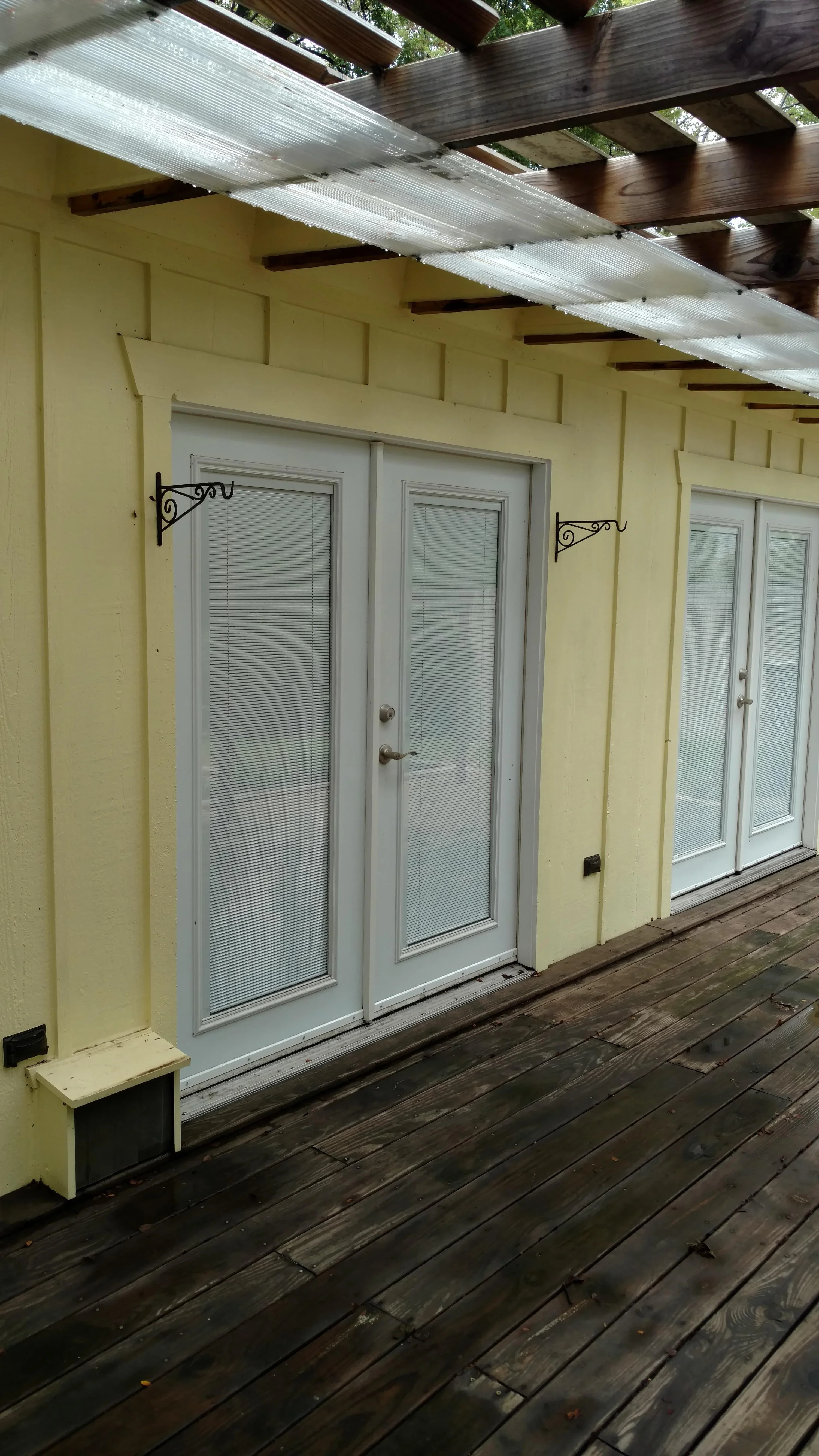 White aluminum patio doors with blinds installed on yellow wall beneath wooden pergola with polycarbonate roof panels