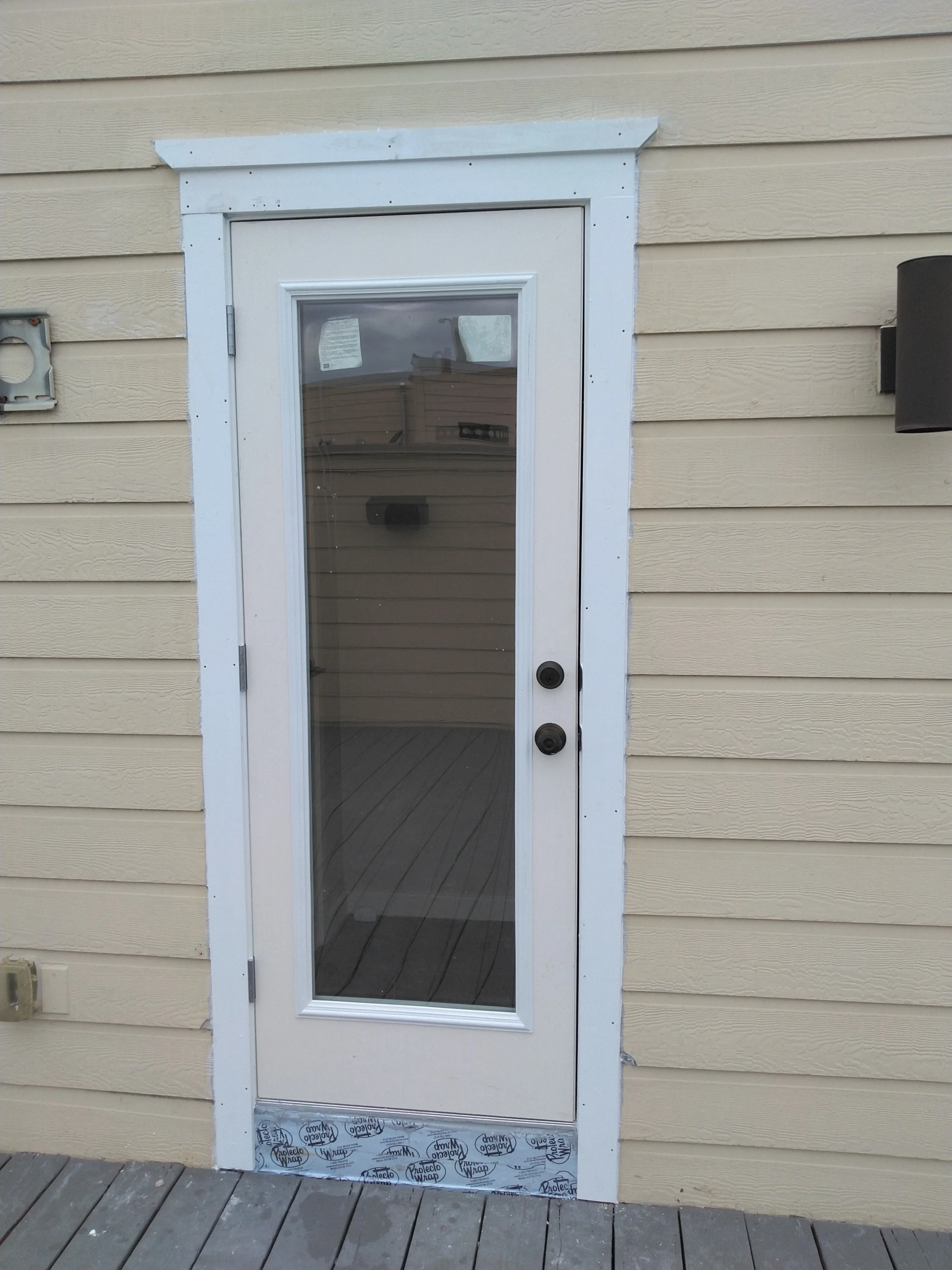 White entry door with glass panel installed in tan vinyl siding, black hardware and threshold visible