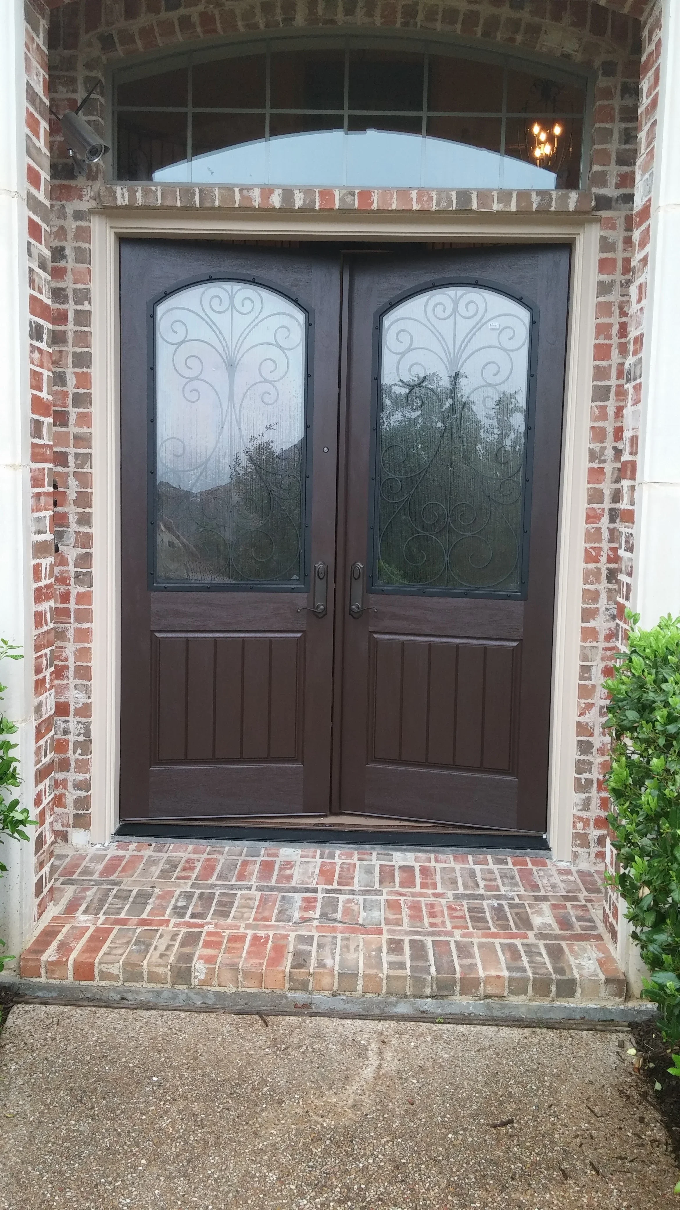 Dark wood double doors with ornamental iron scrollwork glass panels and arched transom window in brick archway