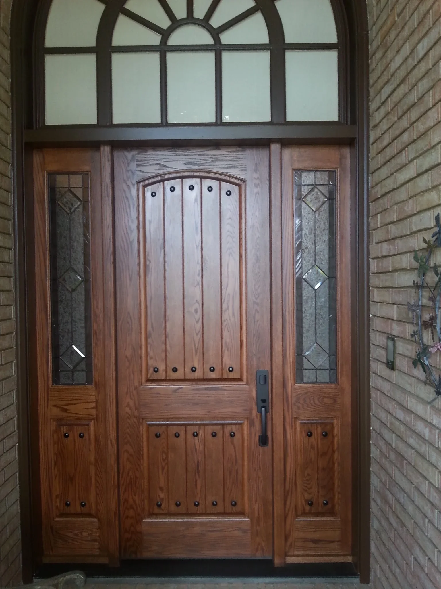 Solid wood entry door with vertical planks, decorative nails, sidelights, and arched transom window
